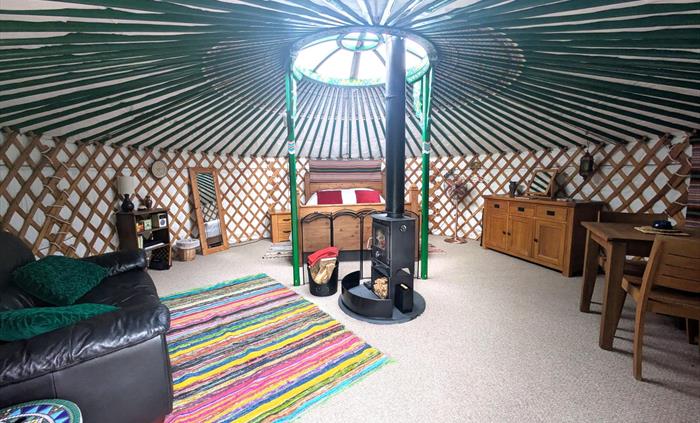 The yurt interior is round with many green painted rafters. There is wood bed at the back, wood table, chairs and cupboard to the right, a black leath